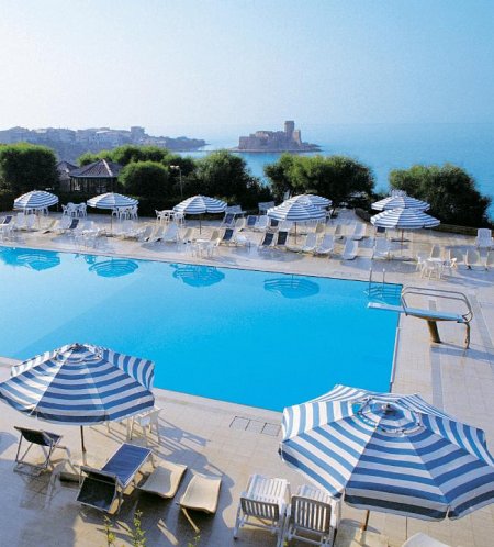 Piscina all'aperto con ombrelloni blu e bianchi, vista mare e un castello in lontananza.