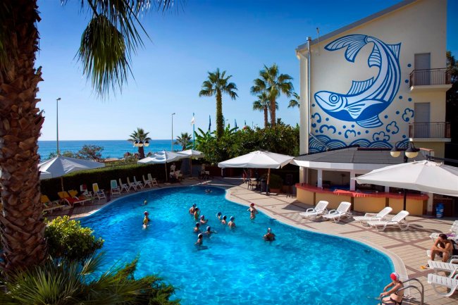 Piscina all'aperto con bagnanti, vista del mare e murale di un pesce blu sull'edificio.