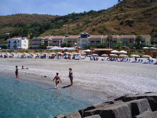 Persone giocano in acqua su una spiaggia di ciottoli, case e colline sullo sfondo.