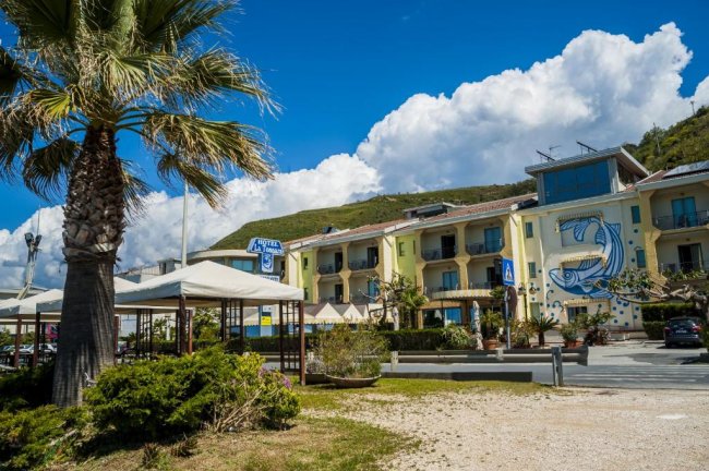Hotel giallo con murale blu, palme e tettoie davanti; cielo blu con nuvole.