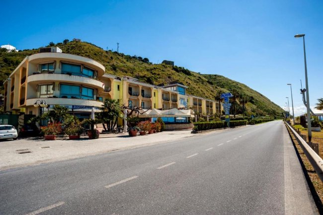 Edificio hotel giallo con balconi curvi, situato lungo una strada con sfondo montuoso.