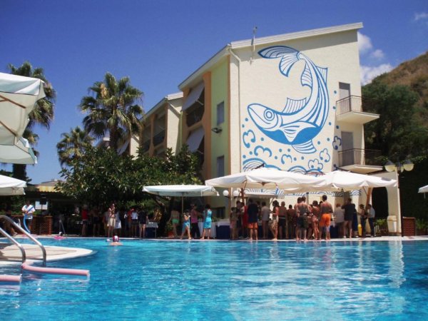 Piscina con persone, edificio con murale di un pesce blu su sfondo bianco.