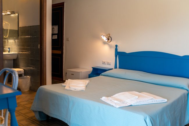 Camera con letto matrimoniale blu, bagno in vista, lampada da parete e asciugamani piegati.