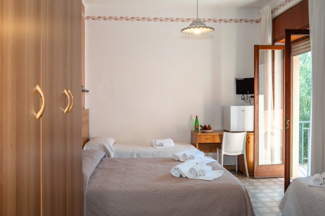 Camera con tre letti, armadio in legno, scrivania, TV, mini-frigo e porta finestra.