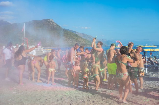 Persone festeggiano sulla spiaggia lanciando polveri colorate.