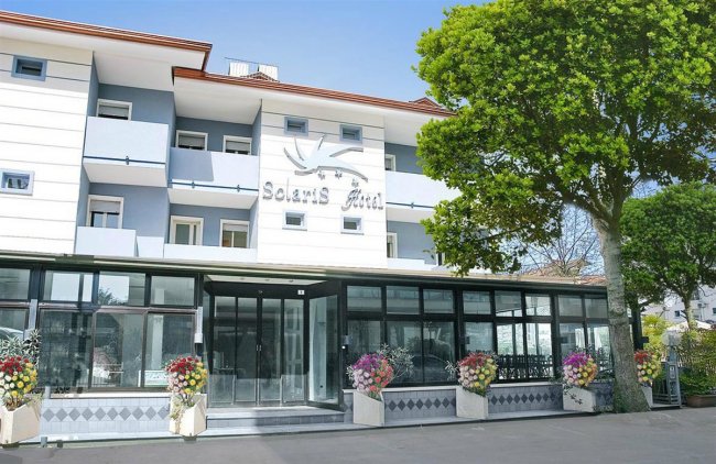 Facciata di un hotel con vetrate, fioriere e un grande albero a destra.